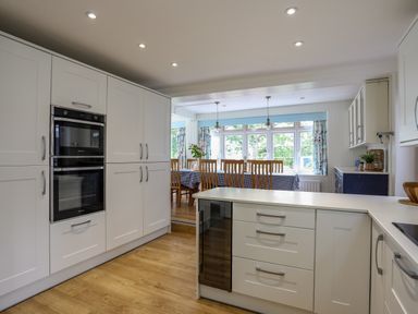 A kitchen with white cabinets and built in ovens next to a dining area with a long table and wooden chairs at Ynys Castell Menai Bridge
