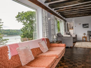 A living room with sofas and armchairs by large windows overlooking trees and water at Ynys Castell in Menai Bridge