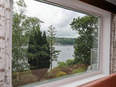 A window view of trees and a lake with a sailboat beyond greenery at Ynys Castell in Menai Bridge