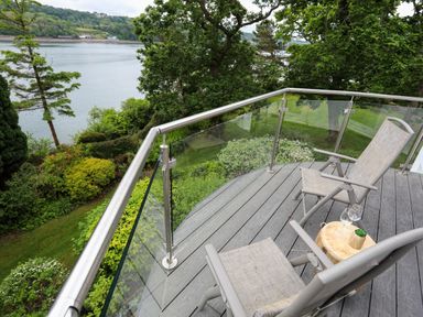 A balcony with two chairs a small table with glasses and a plant overlooking trees and water at Ynys Castell in Menai Bridge