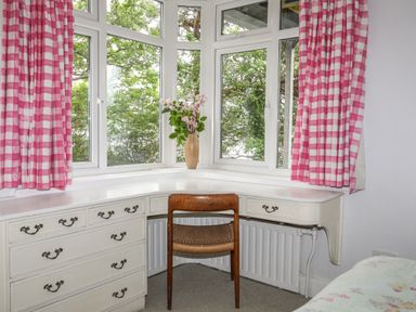 A corner desk with drawers and a wooden chair by windows with pink check curtains and a vase with flowers at Ynys Castell in Menai Bridge