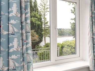 A window with blue curtains showing sailboats and lighthouses with a view of trees shrubs and water outside at Ynys Castell in Menai Bridge