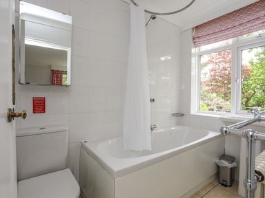 A bathroom with a bathtub and shower curtain a toilet with a mirror above it a window with a pink patterned blind and a small trash bin Ynys Castell Menai Bridge