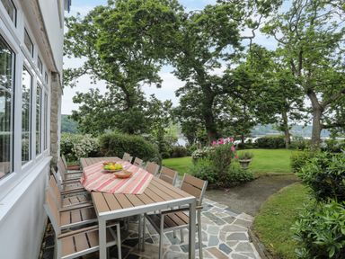 An outdoor patio dining area with a table and chairs next to a garden with trees and flowers at Ynys Castell in Menai Bridge