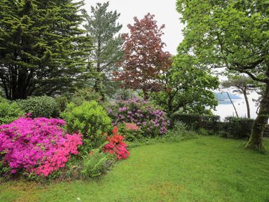 A garden with green grass colorful flowering bushes trees and a body of water in the background at Ynys Castell in Menai Bridge