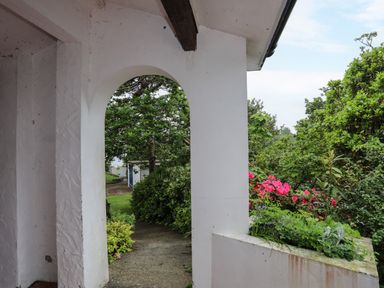 A white arched porch overlooking a garden path with green trees and pink flowers at Ynys Castell in Menai Bridge