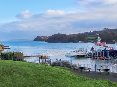 A waterfront area with a grassy foreground two benches a railing a dock with a bridge and boats in the water at Ynys Castell in Menai Bridge