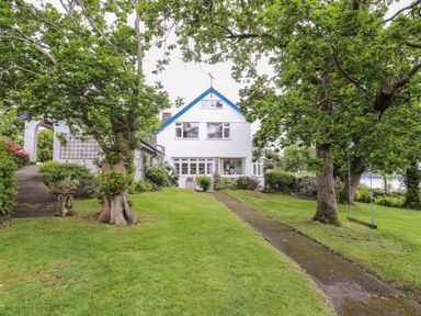 A house with white walls and blue trim surrounded by grass and trees with a stone pathway leading to it at Ynys Castell Menai Bridge