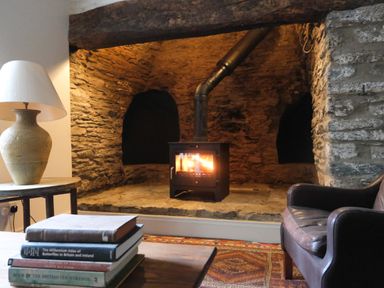 A fireplace with a burning stove in a stone hearth a brown leather chair a stack of books on a wooden table and a table lamp at Henllys Newport Pembrokeshire