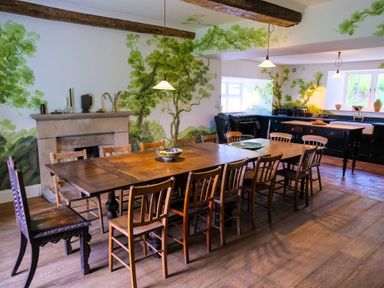 A dining room with a long wooden table, multiple chairs, a fireplace, and a kitchen area with windows at Henllys Newport Pembrokeshire