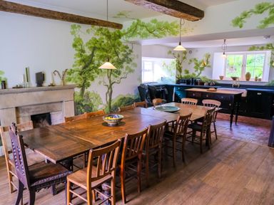 A kitchen dining room with a long wooden dining table and chairs a fireplace and green tree wall murals at Henllys in Newport Pembrokeshire