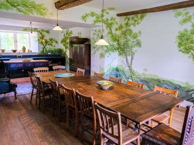 A dining room with a long wooden table and wooden chairs with a kitchen area in the background with a sink and window and painted tree murals on the walls at Henllys in Newport Pembrokeshire