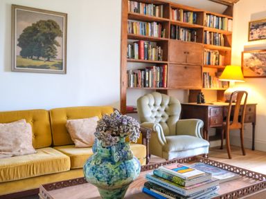 A living room with a yellow sofa, a green armchair, a wooden desk with a lamp, a bookshelf filled with books, and a coffee table with a blue vase and stacked books at Henllys in Newport Pembrokeshire