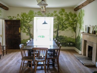 A dining room with a wooden table and chairs with a painted tree mural on the walls and a stone fireplace at Henllys in Newport Pembrokeshire