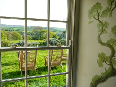 A window view showing outdoor wicker chairs on grass with trees and hills in the background at Henllys in Newport Pembrokeshire