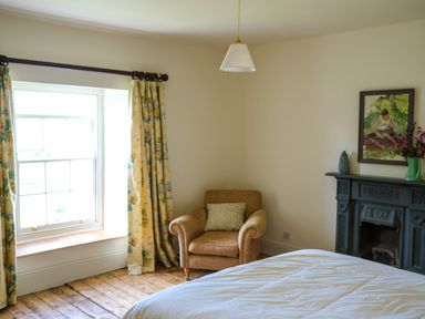 A bedroom with a bed a brown armchair by a window with floral curtains and a dark fireplace with a painting at Henllys in Newport Pembrokeshire