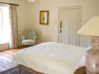 A bedroom with a white bedspread an armchair curtains a framed picture a door and a lamp on a table at Henllys in Newport Pembrokeshire