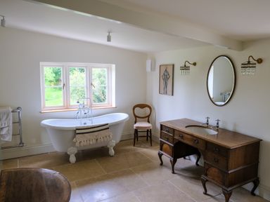 A bathroom with a clawfoot tub by a window a wooden chair a wooden vanity with a sink and an oval mirror at Henllys Newport Pembrokeshire