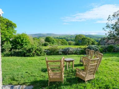 Four wicker chairs and a small wooden table on a grass lawn with trees and hills in the background at Henllys Newport Pembrokeshire