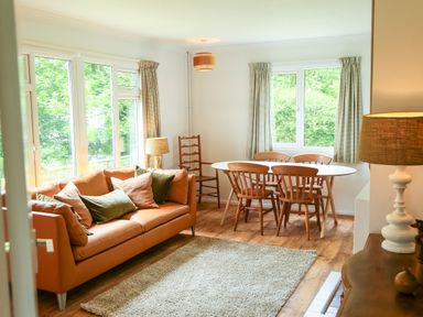 A living room with a brown sofa with cushions a white oval dining table with wooden chairs a floor lamp and windows with curtains at Henllys Lodge in Newport Pembrokeshire
