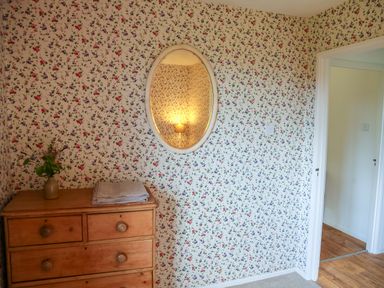 A wooden dresser with a vase and folded cloths beneath an oval mirror on a floral wallpapered wall at Henllys Lodge in Newport Pembrokeshire