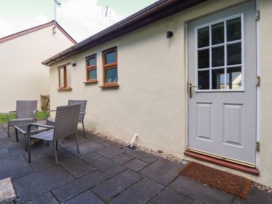 An outdoor patio area with four wicker chairs and a glass table beside a cream house with three small windows and a white door at Lilyvale in Davidstow near Camelford