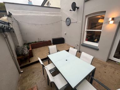An outdoor patio with a glass table and six chairs a small raised brick planter a wall clock a satellite dish and string lights at Green Retreat in Dartmouth