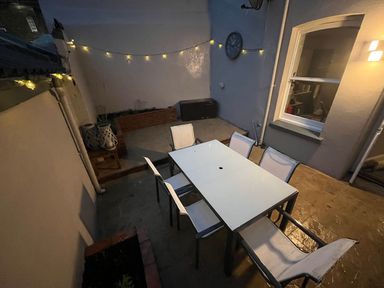 An outdoor patio area with a glass dining table and six chairs fairy lights on walls and a clock at Green Retreat in Dartmouth