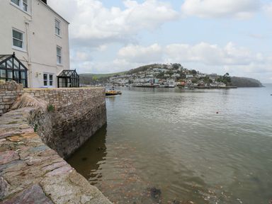 A waterfront stone wall with a building on the left and a marina with houses on a hill in the background at Green Retreat in Dartmouth