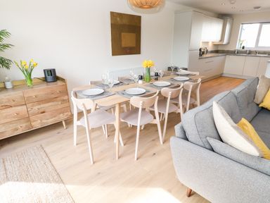 A dining area with a wooden table set for eight people next to a gray sofa and white kitchen cabinets at The Bright House in St Eval near St Columb Major
