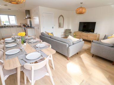An open plan living and dining area with a wooden dining table set for six and grey sofas facing a television at The Bright House in St Eval near St Columb Major