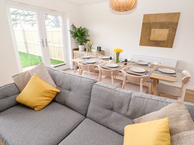 A dining area with a wooden table set for six and a grey couch with yellow and beige cushions at The Bright House in St Eval near St Columb Major