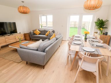 A living room and dining area with grey sofas yellow cushions wooden dining table set with plates and glasses at The Bright House in St Eval near St Columb Major