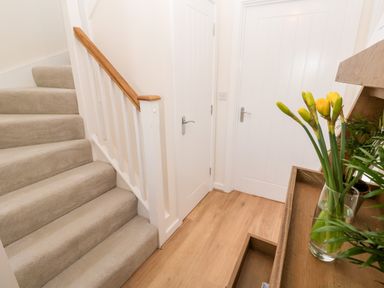 A staircase with beige carpet and wooden handrail next to two white doors and a wooden table with yellow flowers and green plants at The Bright House in St Eval near St Columb Major
