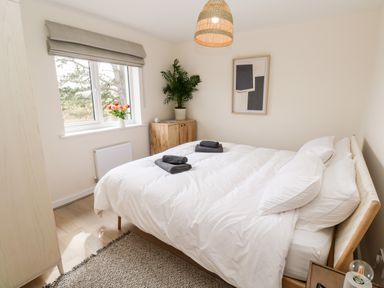 A bedroom with a bed with white bedding two gray towels a wooden cabinet a plant a window with flowers and a framed picture at The Bright House in St Eval near St Columb Major