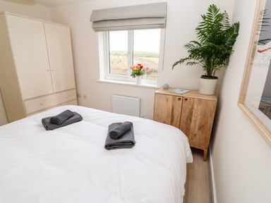 A bedroom with a bed with white sheets and gray towels by a window with flowers a wooden cabinet with a plant and wardrobe at The Bright House in St Eval near St Columb Major