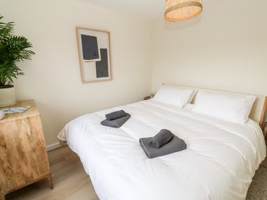 A bedroom with a double bed white bedding and folded towels on the bed a wooden chest of drawers with a plant and framed art on the wall at The Bright House in St Eval near St Columb Major