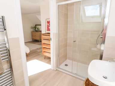 A bathroom with a glass shower enclosure a white sink and wooden drawers visible through the open door at The Bright House in St Eval near St Columb Major