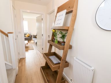 A hallway with a wooden ladder-style shelf holding plants and a picture a round wall clock and a radiator at The Bright House in St Eval near St Columb Major