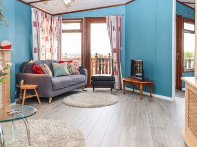 A living room with a blue sofa and colorful cushions a small wooden table with a lamp a TV on a wooden corner table and sliding doors with curtains at Lodge Two Country View Park Graveney