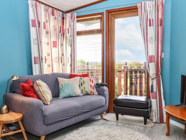 A small living room with a blue sofa and colorful cushions next to a glass door and windows with patterned curtains at Lodge Two Country View Park in Graveney