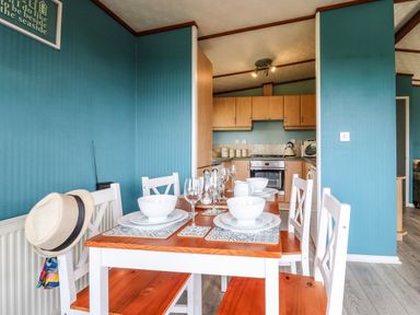 A dining area with a wooden table set with white bowls and glasses below a blue wall with a kitchen in the background at Lodge Two Country View Park in Graveney