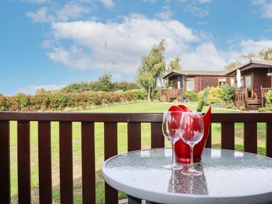 A glass table with two wine glasses and a bottle in a red container on a wooden deck outside two wooden lodges in Graveney