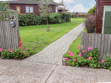 A paved pathway between grass and pink flowers leading to wooden buildings at Lodge Two Country View Park in Graveney