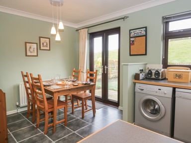 A dining area with a wooden table and chairs near glass doors and a kitchen counter with a washing machine and toaster at Marsh Garth in Kirkby-In-Furness