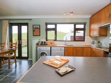 A kitchen with wooden cabinets a countertop with cheese and crackers a dining table and glass doors at Marsh Garth in Kirkby-In-Furness