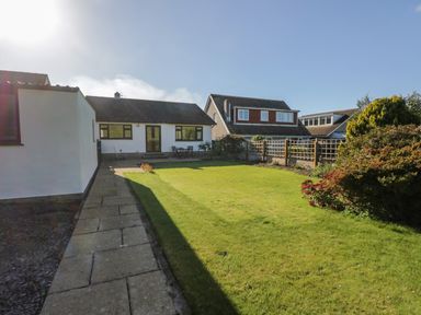 A backyard with a paved pathway lawn and bushes and a white house with black framed windows at Marsh Garth in Kirkby-In-Furness