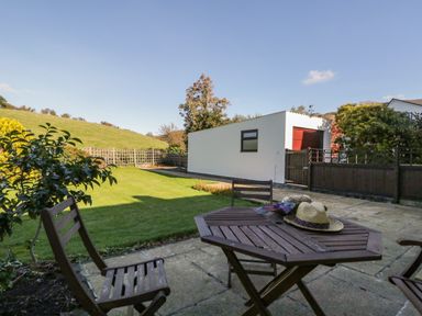 A patio with wooden chairs and table with hats on it and a lawn with a white building in the background at Marsh Garth in Kirkby-In-Furness