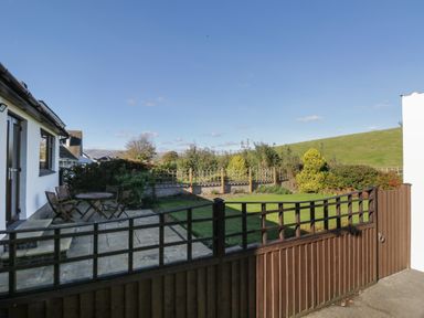 A backyard with a stone patio featuring a wooden table and chairs a grassy area with shrubs and a wooden fence under a clear sky at Marsh Garth in Kirkby-In-Furness