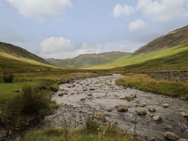 A river with rocks flowing between grassy hills and a stone wall under a partly cloudy sky at Marsh Garth in Kirkby-In-Furness